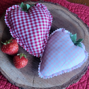 Pu&ograve; includere: Due decorazioni in tessuto a forma di cuore con motivi a quadretti, una rossa e bianca, l'altra rosa e bianca, sono appoggiate su una fetta di legno. Sono presenti anche due fragole fresche, che suggeriscono un tema di fascino rustico e artigianato.