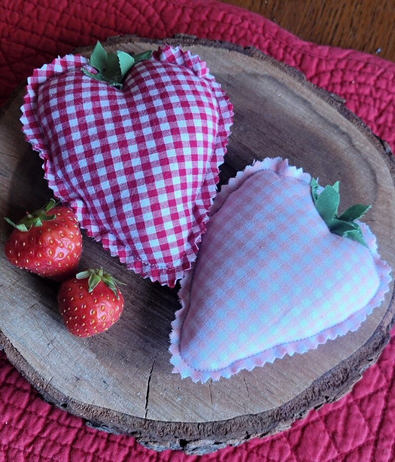 Pu&ograve; includere: Due decorazioni in tessuto a forma di cuore con motivi a quadretti, una rossa e bianca, l'altra rosa e bianca, sono appoggiate su una fetta di legno. Sono presenti anche due fragole fresche, che suggeriscono un tema di fascino rustico e artigianato.