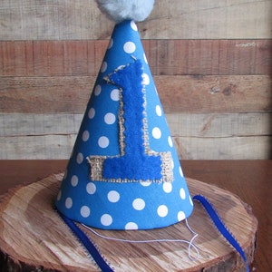 May include: A conical party hat in bright blue with white polka dots, featuring a large blue felt number one appliqued onto a burlap patch on the front. The top is finished with a fluffy light blue pom-pom, and thin blue ribbons are attached.