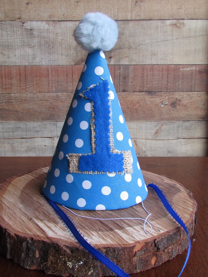 May include: A conical party hat in bright blue with white polka dots, featuring a large blue felt number one appliqued onto a burlap patch on the front. The top is finished with a fluffy light blue pom-pom, and thin blue ribbons are attached.