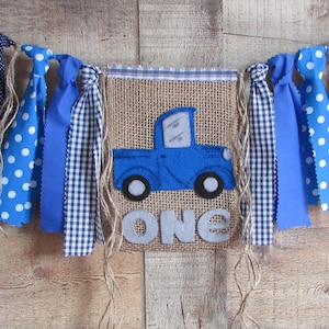 May include: A burlap banner featuring a blue felt truck appliqu&eacute; and the word ONE in grey felt letters. The banner is decorated with hanging fabric strips in blue and white polka dot and gingham patterns, tied with twine to a top fabric border.
