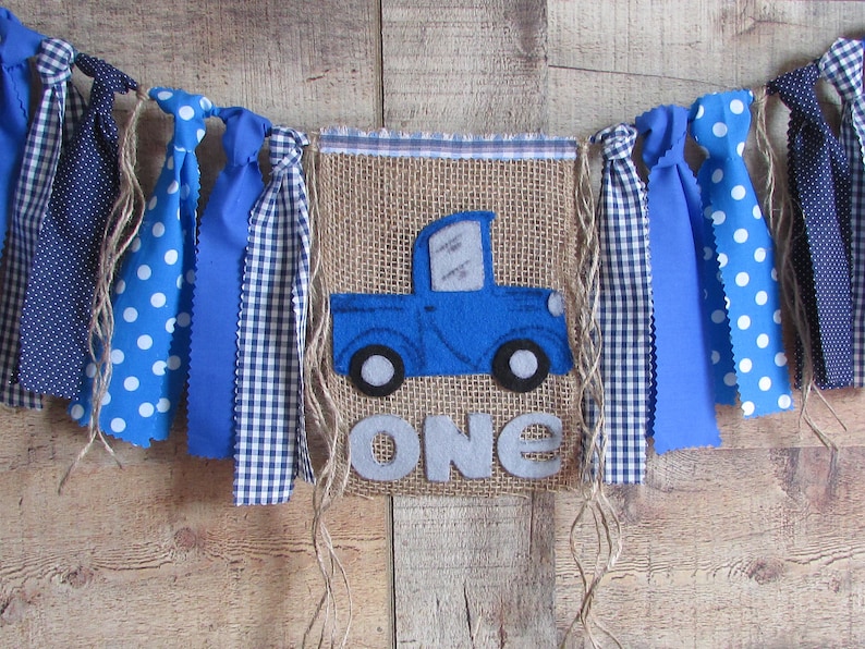 May include: A burlap banner featuring a blue felt truck appliqu&eacute; and the word ONE in grey felt letters. The banner is decorated with hanging fabric strips in blue and white polka dot and gingham patterns, tied with twine to a top fabric border.