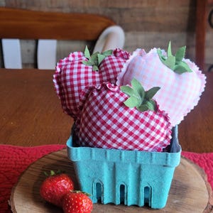 Pu&ograve; includere: Una natura morta rustica presenta un cestino di bacche azzurro chiaro pieno di fragole di stoffa a forma di cuore con motivi a quadri rossi e bianchi e rosa. Fragole fresche poggiano su una fetta di legno.