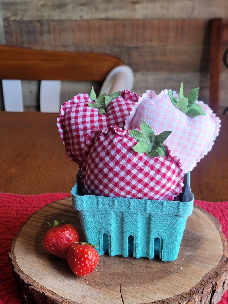 Pu&ograve; includere: Una natura morta rustica presenta un cestino di bacche azzurro chiaro pieno di fragole di stoffa a forma di cuore con motivi a quadri rossi e bianchi e rosa. Fragole fresche poggiano su una fetta di legno.
