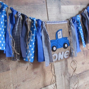 May include: A decorative fabric banner featuring a central burlap pennant with a blue truck applique and the word ONE in grey felt letters. The banner is adorned with various blue and white patterned fabric strips and hanging twine strands.