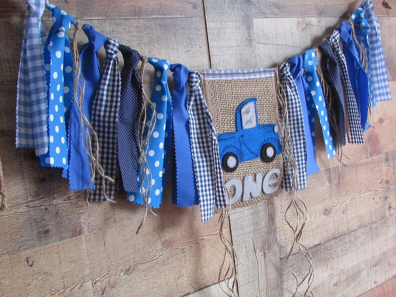 May include: A decorative fabric banner featuring a central burlap pennant with a blue truck applique and the word ONE in grey felt letters. The banner is adorned with various blue and white patterned fabric strips and hanging twine strands.