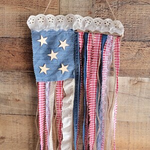 K&ouml;nnte beinhalten: Eine patriotische Wanddekoration in Form der amerikanischen Flagge. Sie hat einen blauen Denim-Bereich mit Holzsternen, rot-wei&szlig;em Vichy, Spitze und Denim-Streifen. Der obere Rand hat eine wei&szlig;e Lochspitze und wird an einer Schnur aufgeh&auml;ngt.