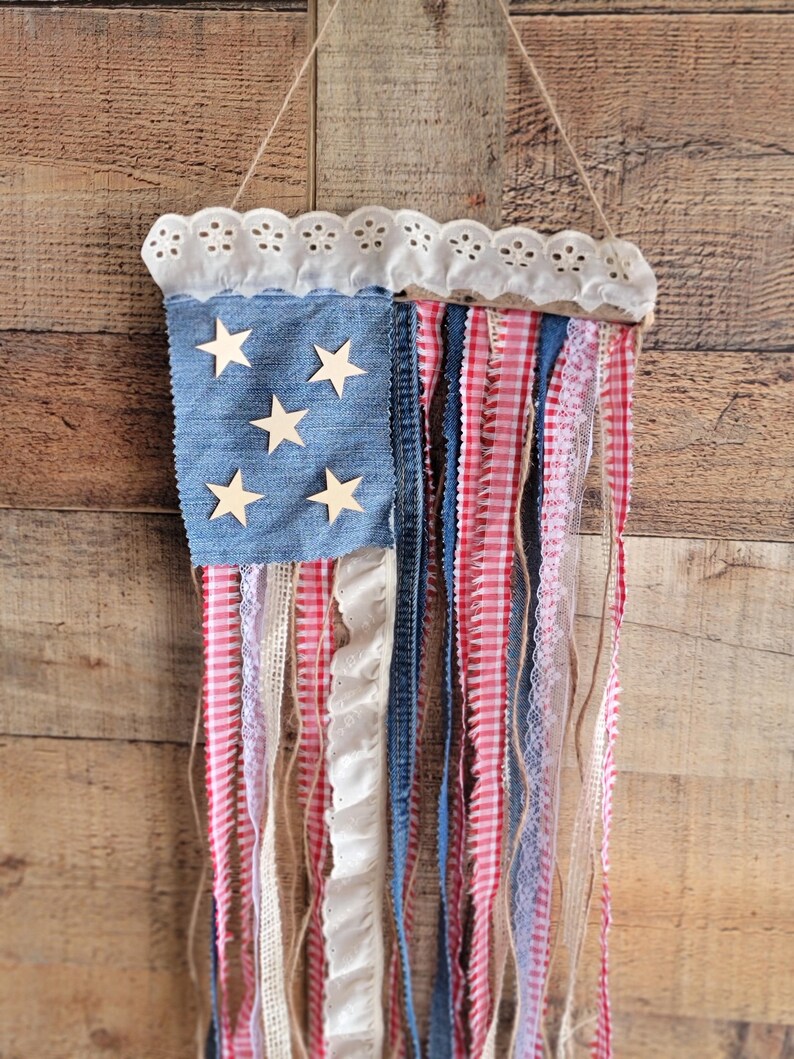 K&ouml;nnte beinhalten: Eine patriotische Wanddekoration in Form der amerikanischen Flagge. Sie hat einen blauen Denim-Bereich mit Holzsternen, rot-wei&szlig;em Vichy, Spitze und Denim-Streifen. Der obere Rand hat eine wei&szlig;e Lochspitze und wird an einer Schnur aufgeh&auml;ngt.