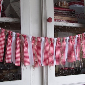 May include: A festive red and white gingham fabric garland with white fabric strips and twine accents. The garland is strung across a white cabinet with chicken wire detailing, evoking a rustic, country aesthetic. The garland is perfect for home decor.