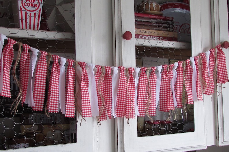 May include: A festive red and white gingham fabric garland with white fabric strips and twine accents. The garland is strung across a white cabinet with chicken wire detailing, evoking a rustic, country aesthetic. The garland is perfect for home decor.