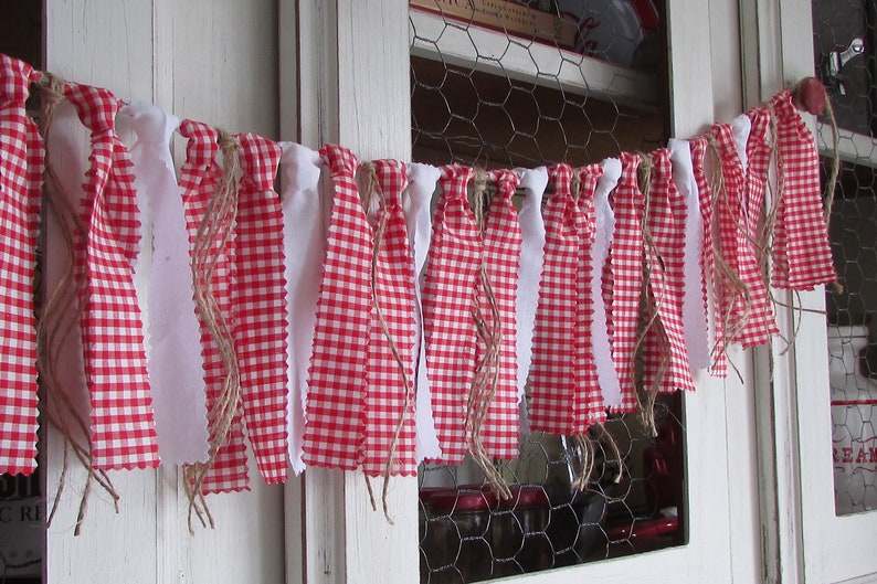 May include: A decorative banner featuring alternating red and white gingham and solid white fabric strips. The strips are attached to a natural twine, creating a rustic, country-style aesthetic. The banner is suitable for home decor.