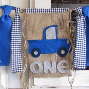 May include: A burlap banner featuring a blue felt truck applique and the word ONE in grey felt letters. The banner is topped with a blue and white gingham trim and decorated with hanging fabric strips in blue polka dot and blue gingham patterns.