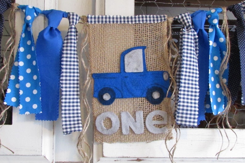 May include: A burlap banner featuring a blue felt truck applique and the word ONE in grey felt letters. The banner is topped with a blue and white gingham trim and decorated with hanging fabric strips in blue polka dot and blue gingham patterns.