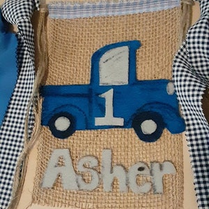 May include: A rectangular burlap banner featuring a blue felt truck appliqu&eacute; with the number 1 on the side and the name Asher in white felt letters below. The banner is trimmed with a blue and white striped fabric top and black and white gingham ribbons.