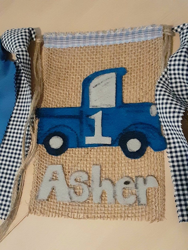 May include: A rectangular burlap banner featuring a blue felt truck appliqu&eacute; with the number 1 on the side and the name Asher in white felt letters below. The banner is trimmed with a blue and white striped fabric top and black and white gingham ribbons.