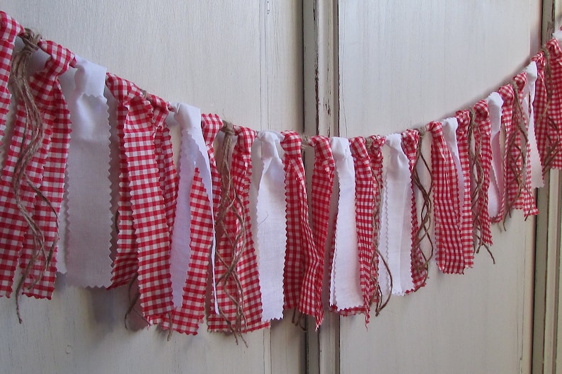 May include: A decorative banner featuring alternating red and white gingham fabric strips and solid white fabric strips. The banner is strung together with twine, creating a rustic and charming aesthetic. Perfect for festive occasions.