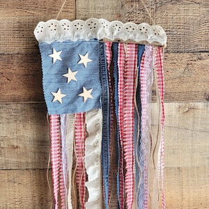 K&ouml;nnte beinhalten: Eine patriotische Wanddekoration in Form der amerikanischen Flagge. Sie hat einen jeansblauen Abschnitt mit Holzsternen und rot-wei&szlig; gestreiften Stoffstreifen. Die Oberseite hat eine wei&szlig;e Spitzentrimmung, und die Aufh&auml;ngung ist aus Bindfaden.