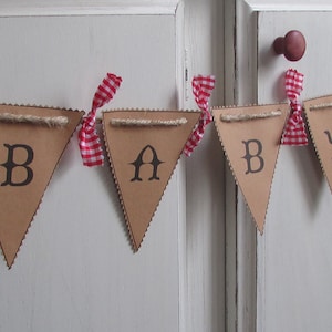 May include: A brown cardboard banner spelling "BABY" with black letters. Each pennant is trimmed with a decorative edge and strung with twine. Red and white gingham ribbons are tied between each pennant, creating a festive look.