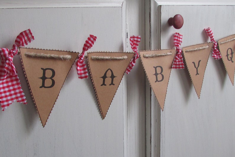 May include: A brown cardboard banner spelling "BABY" with black letters. Each pennant is trimmed with a decorative edge and strung with twine. Red and white gingham ribbons are tied between each pennant, creating a festive look.