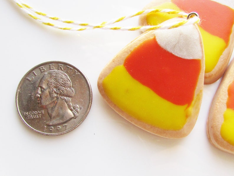 Puede incluir: Tres galletas con forma de ma&iacute;z dulce amarillo, naranja y blanco con una cuerda blanca atada a la parte superior.