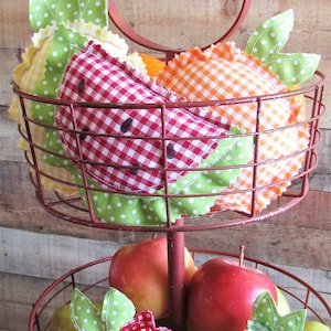 May include: A two-tiered red wire basket containing fabric fruit ornaments and real fruit. The fabric ornaments feature red, orange, and yellow gingham patterns with green polka-dot leaves. The basket holds a red watermelon slice, orange, and apple shapes.