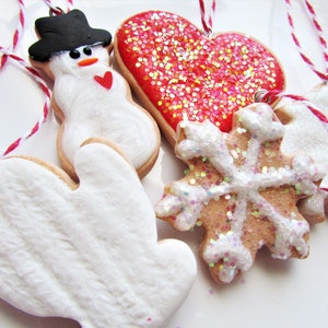May include: Five Christmas ornaments made of gingerbread cookies. The ornaments are decorated with white icing and glitter. The ornaments are shaped like a snowman, a heart, a snowflake, a mitten, and a Christmas tree.