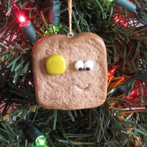 May include: A brown clay ornament shaped like a slice of toast with a yellow circle representing butter and googly eyes. The ornament is hanging on a green artificial Christmas tree.
