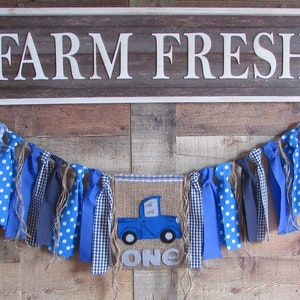 May include: A rectangular sign with the white text FARM FRESH and a fabric banner featuring a blue truck graphic, the word ONE, and various blue and white patterned fabric strips tied to a jute twine string for decorative display.