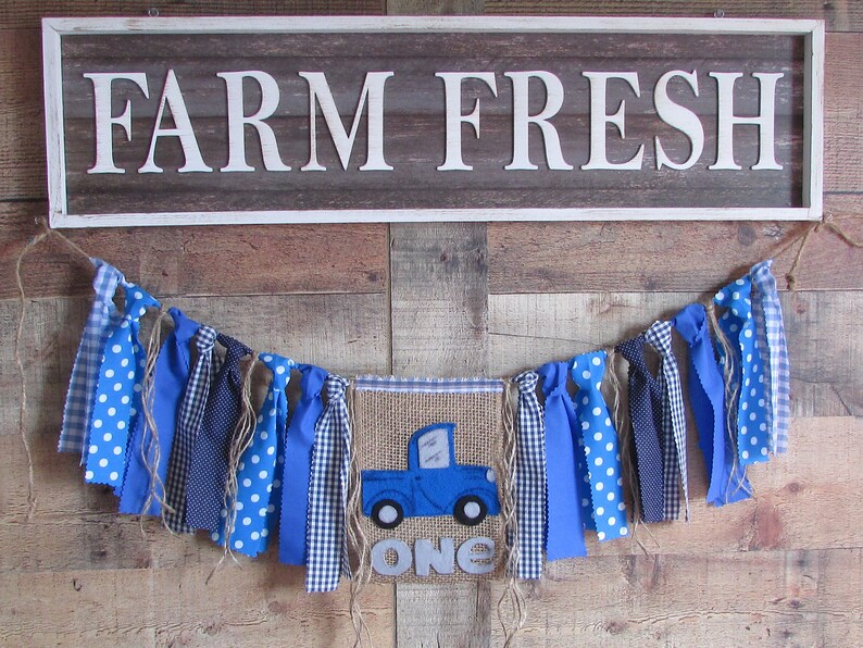 May include: A rectangular sign with the white text FARM FRESH and a fabric banner featuring a blue truck graphic, the word ONE, and various blue and white patterned fabric strips tied to a jute twine string for decorative display.