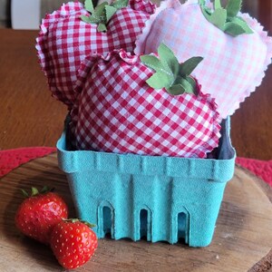 Pu&ograve; includere: Un cestino di bacche azzurro chiaro pieno di fragole di stoffa. Le fragole sono a quadretti rossi e bianchi e a quadretti rosa con foglie di stoffa verdi. Due fragole rosse fresche sono davanti al cestino su una superficie di legno.