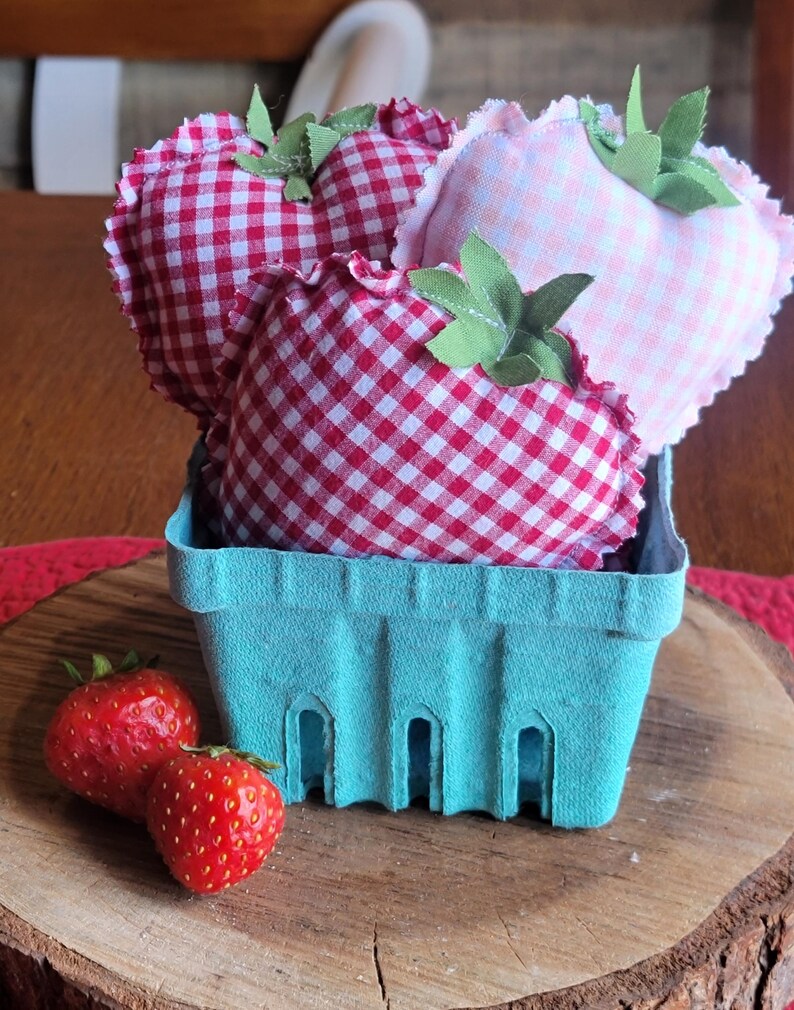 Pu&ograve; includere: Un cestino di bacche azzurro chiaro pieno di fragole di stoffa. Le fragole sono a quadretti rossi e bianchi e a quadretti rosa con foglie di stoffa verdi. Due fragole rosse fresche sono davanti al cestino su una superficie di legno.