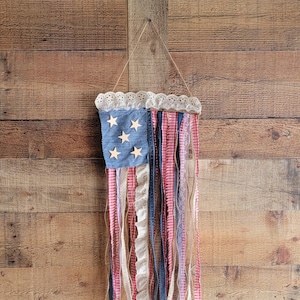 K&ouml;nnte beinhalten: Eine patriotische Wanddekoration in Form der amerikanischen Flagge. Sie hat einen blauen Denim-Bereich mit Holzsternen und roten, wei&szlig;en und blauen Stoffstreifen. Die Oberseite ist mit wei&szlig;er Spitze verziert.