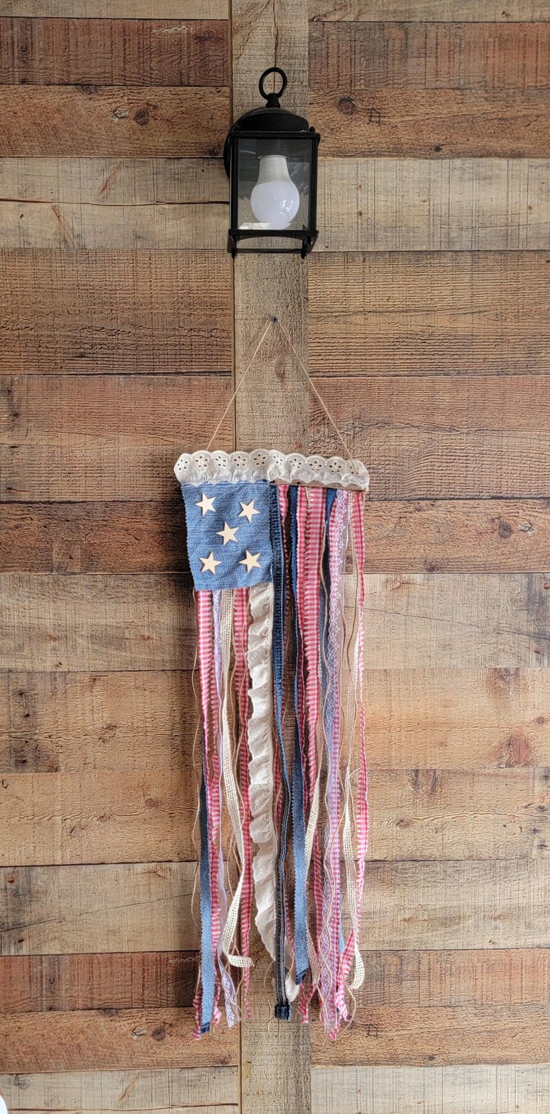 K&ouml;nnte beinhalten: Eine patriotische Wanddekoration in Form der amerikanischen Flagge. Sie hat einen blauen Denim-Bereich mit Holzsternen und roten, wei&szlig;en und blauen Stoffstreifen. Die Oberseite ist mit wei&szlig;er Spitze verziert.