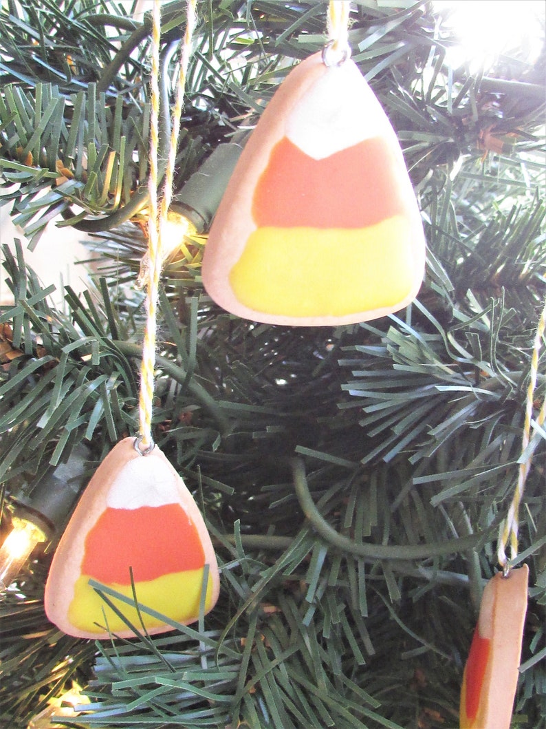 Puede incluir: Tres adornos de ma&iacute;z dulce hechos a mano colgando de un &aacute;rbol de Navidad artificial verde. Los adornos est&aacute;n hechos de arcilla blanca, naranja y amarilla y tienen forma de ma&iacute;z dulce.