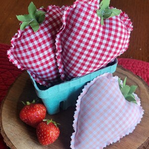 Pu&ograve; includere: Decorazioni a forma di fragola in tessuto, presentate in un cestino azzurro, con fragole fresche su una superficie di legno. Le fragole in tessuto presentano un motivo a quadretti rossi e bianchi, dettagli di foglie verdi e un bordo arricciato.
