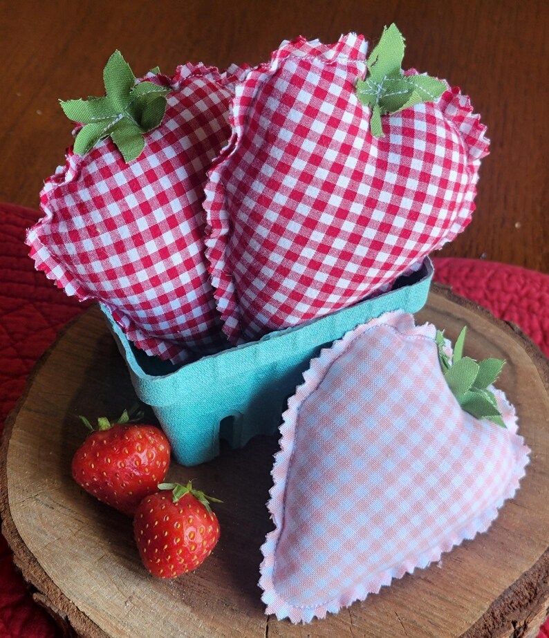 Pu&ograve; includere: Decorazioni a forma di fragola in tessuto, presentate in un cestino azzurro, con fragole fresche su una superficie di legno. Le fragole in tessuto presentano un motivo a quadretti rossi e bianchi, dettagli di foglie verdi e un bordo arricciato.