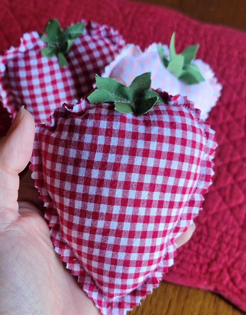 Pu&ograve; includere: Tre decorazioni in tessuto a forma di fragola. Le fragole sono realizzate in tessuto a quadretti rosso e bianco con un bordo a balze. Ognuna ha foglie di tessuto verde attaccate in alto. Lo sfondo &egrave; un tessuto rosso.