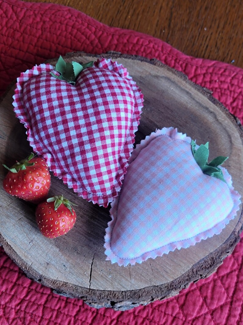 Pu&ograve; includere: Due decorazioni in tessuto a forma di cuore con motivi a quadretti, una rossa e bianca, l'altra rosa e bianca, sono appoggiate su una fetta di legno. Sono presenti anche due fragole fresche. Gli oggetti poggiano su una superficie trapuntata rossa.