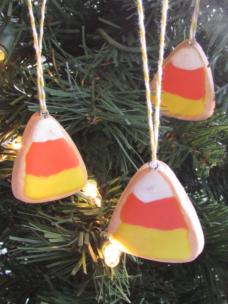 Puede incluir: Tres adornos de ma&iacute;z dulce hechos a mano colgando de un &aacute;rbol de Navidad verde. Los adornos est&aacute;n hechos de arcilla polim&eacute;rica y est&aacute;n pintados en los colores tradicionales del ma&iacute;z dulce, naranja, amarillo y blanco.