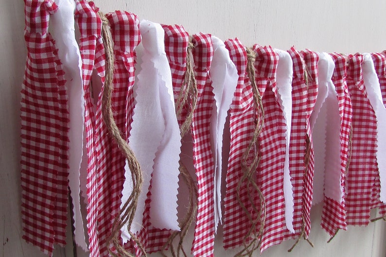 May include: A decorative banner featuring alternating red and white gingham fabric and white fabric strips. The banner is strung on a natural twine rope, creating a rustic and charming aesthetic. Perfect for festive occasions or home decor.