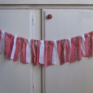 May include: A decorative banner featuring alternating strips of red and white gingham fabric and solid white fabric. The banner is strung on a natural twine, creating a rustic and charming aesthetic. Perfect for festive occasions.