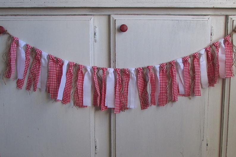 May include: A decorative banner featuring alternating strips of red and white gingham fabric and solid white fabric. The banner is strung on a natural twine, creating a rustic and charming aesthetic. Perfect for festive occasions.