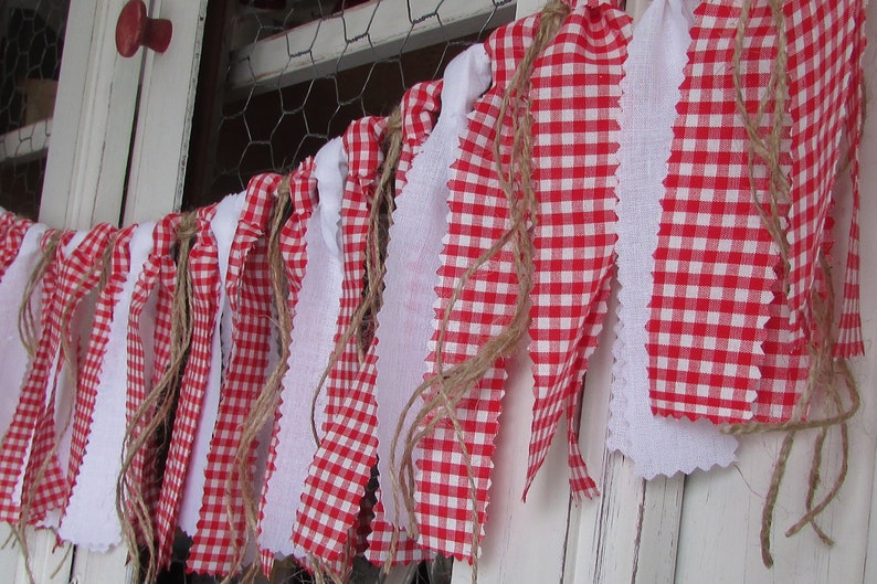 May include: A festive banner with alternating red and white gingham and solid white fabric strips. The edges are raw and frayed, and the banner is strung with natural twine. The banner is hanging in front of a white wooden surface.