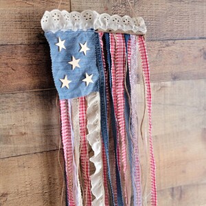 K&ouml;nnte beinhalten: Eine patriotische Wanddekoration, die die amerikanische Flagge darstellt. Sie besteht aus einem quadratischen Denim-Blau mit Holzsternen, rot-wei&szlig;em Vichy, Spitze und Denim-B&auml;ndern. Die Oberseite ist mit wei&szlig;er Spitze verziert und wird von einer Schnur gehalten.