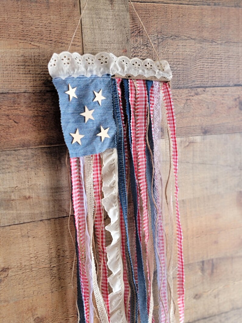 K&ouml;nnte beinhalten: Eine patriotische Wanddekoration, die die amerikanische Flagge darstellt. Sie besteht aus einem quadratischen Denim-Blau mit Holzsternen, rot-wei&szlig;em Vichy, Spitze und Denim-B&auml;ndern. Die Oberseite ist mit wei&szlig;er Spitze verziert und wird von einer Schnur gehalten.