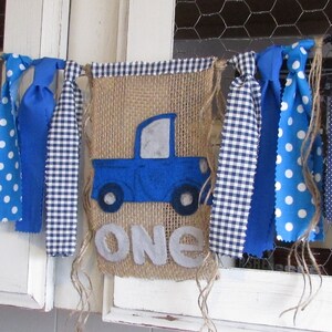 May include: A decorative banner featuring a central burlap panel with a blue felt truck applique and the word ONE in light gray felt letters. The banner is accented with various blue and white fabric strips in polka dot and gingham patterns.