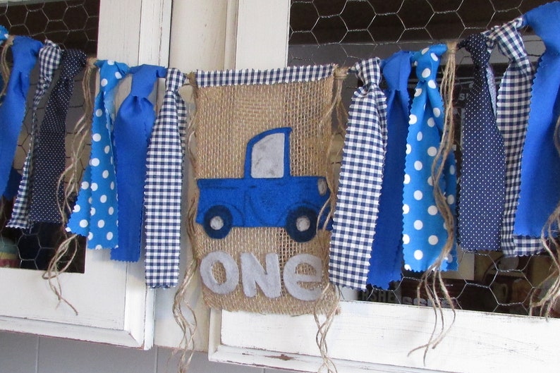 May include: A decorative banner featuring a central burlap panel with a blue felt truck applique and the word ONE in light gray felt letters. The banner is accented with various blue and white fabric strips in polka dot and gingham patterns.