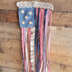 K&ouml;nnte beinhalten: Eine patriotische Wanddekoration, die die amerikanische Flagge darstellt. Sie hat ein denimblaues Quadrat mit Holzsternen, rot-wei&szlig; gestreiften B&auml;ndern und wei&szlig;em Spitzensaum. Der Aufh&auml;nger wird von einem nat&uuml;rlichen Bindfaden gehalten.