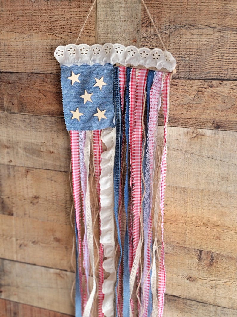K&ouml;nnte beinhalten: Eine patriotische Wanddekoration, die die amerikanische Flagge darstellt. Sie hat ein denimblaues Quadrat mit Holzsternen, rot-wei&szlig; gestreiften B&auml;ndern und wei&szlig;em Spitzensaum. Der Aufh&auml;nger wird von einem nat&uuml;rlichen Bindfaden gehalten.