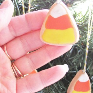 Puede incluir: Una mano sostiene un adorno de ma&iacute;z dulce amarillo, naranja y blanco. El adorno est&aacute; colgado de una cuerda en un &aacute;rbol de Navidad artificial verde.